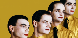 Uma releitura artística da icônica imagem da banda Kraftwerk, apresentando quatro homens em uma composição diagonal. Eles possuem pele muito clara, cabelos escuros penteados para trás e lábios pintados com batom vermelho vibrante. Todos vestem camisas de um tom amarelo-ouro cintilante com gravatas pretas finas, posicionados contra um fundo sólido também na cor amarelo-ouro. O estilo visual remete à estética retrô-futurista e minimalista do grupo original.