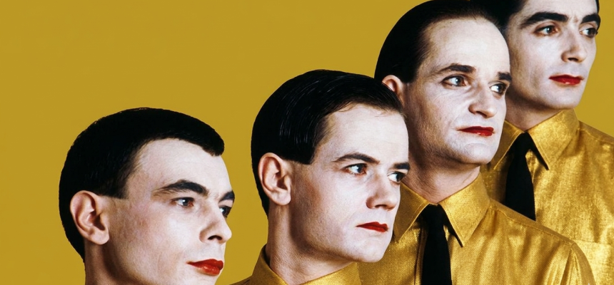 Uma releitura artística da icônica imagem da banda Kraftwerk, apresentando quatro homens em uma composição diagonal. Eles possuem pele muito clara, cabelos escuros penteados para trás e lábios pintados com batom vermelho vibrante. Todos vestem camisas de um tom amarelo-ouro cintilante com gravatas pretas finas, posicionados contra um fundo sólido também na cor amarelo-ouro. O estilo visual remete à estética retrô-futurista e minimalista do grupo original.